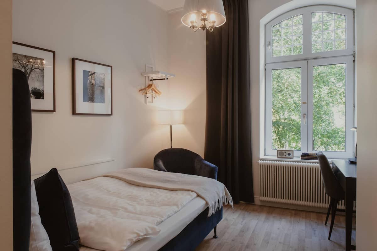 A bedroom with a bed, a desk, a chair, a radiator, a window, and two framed pictures on the wall.
