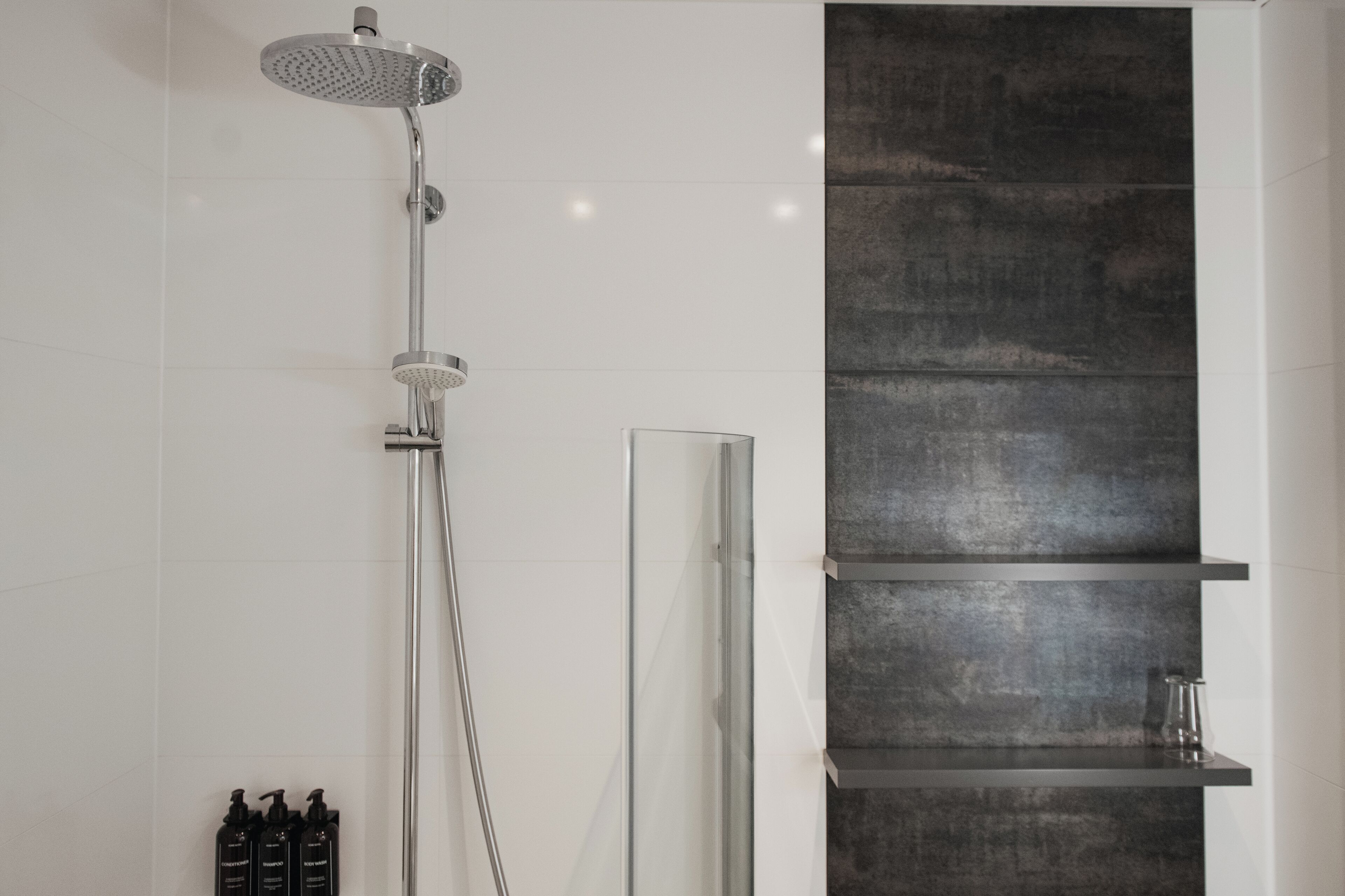 A modern bathroom with a glass-enclosed shower, a vertical dark-textured wall, and a minimalist shelf unit.