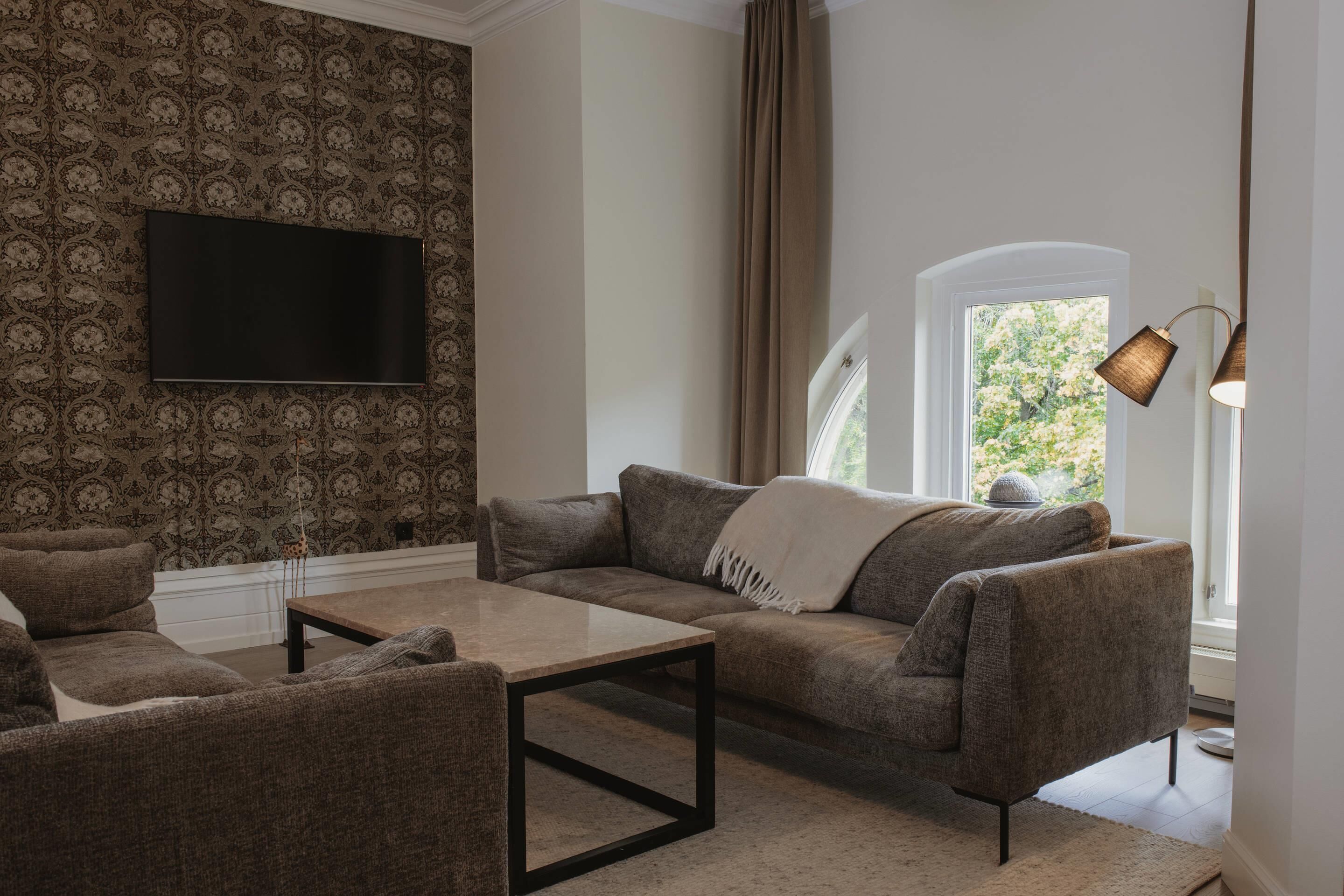 Un salon comprenant un canapé, un fauteuil et une table basse, un téléviseur à écran plat fixé à un mur orné de motifs, et une fenêtre avec des rideaux.
