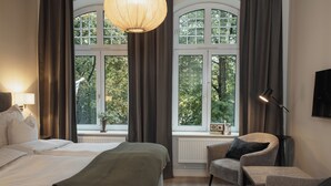 Habitación de hotel con una cama grande, dos sillones, una mesita, una lámpara y vistas a árboles a través de amplios ventanales.