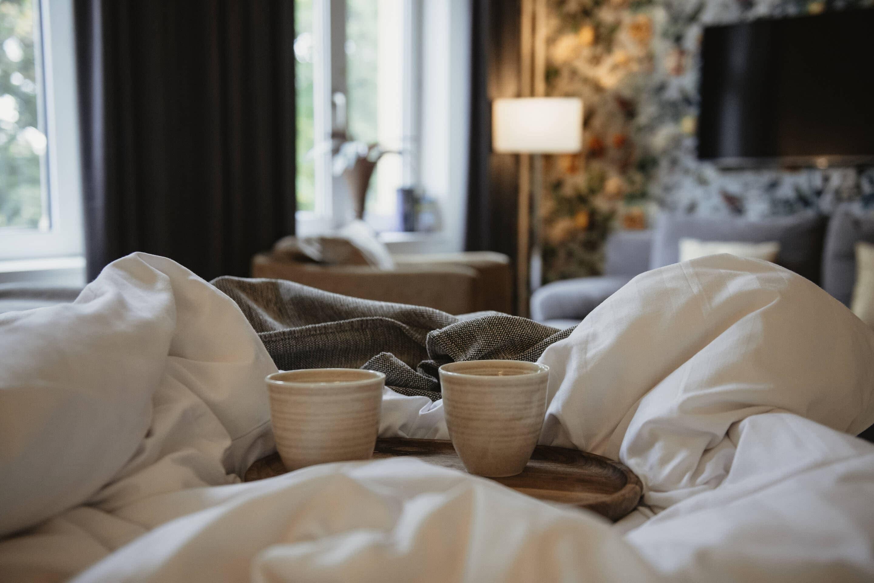 A bed with white linens, a bedside table with a lamp, and a view of a cityscape through the window.