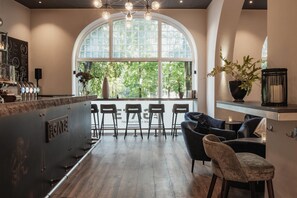 A modern bar area with a large arched window, bar stools, and a seating area with a sofa and chairs. - Home Hotel Norre Park (Halmstad)