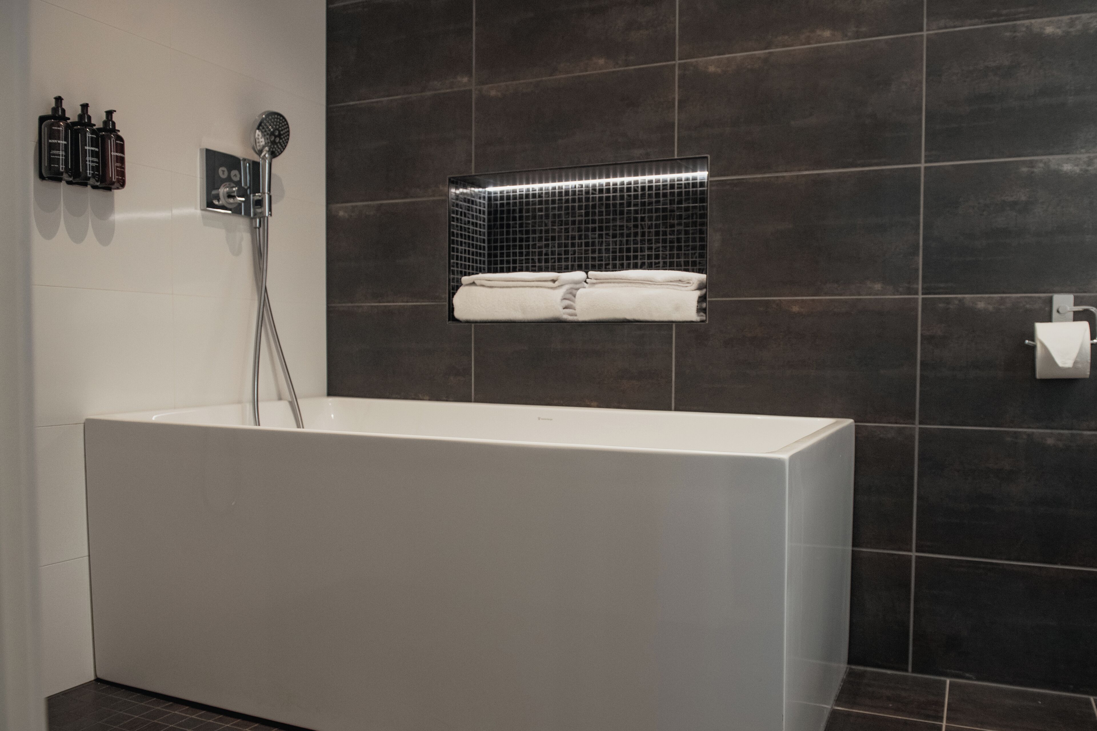 A modern bathroom with a large white bathtub, a handheld showerhead, and a shelf with folded towels.