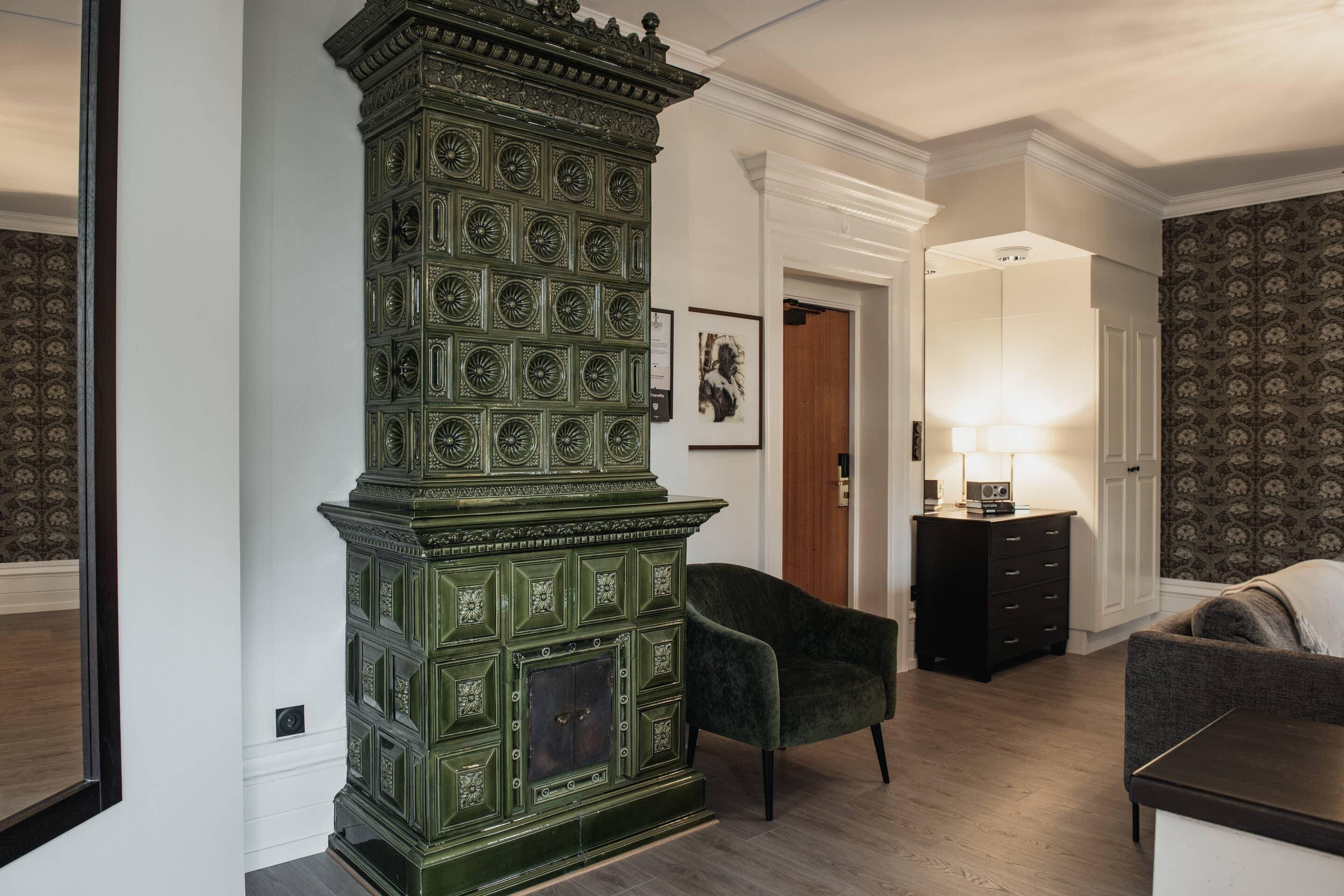 Una habitación con una chimenea verde grande, un aparador de madera oscura, un sillón verde y un papel tapiz estampado.