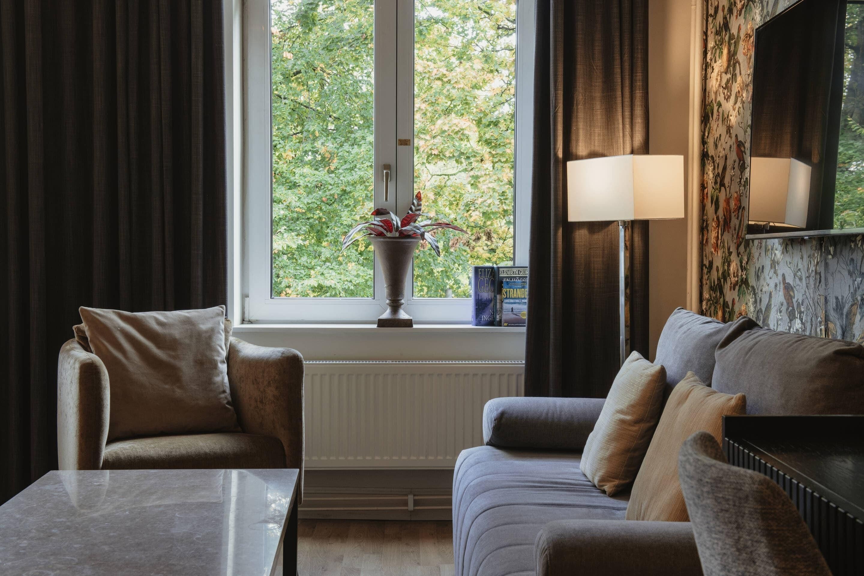 A modern living room with a sofa, a coffee table, a potted plant, and a window with a view of trees.