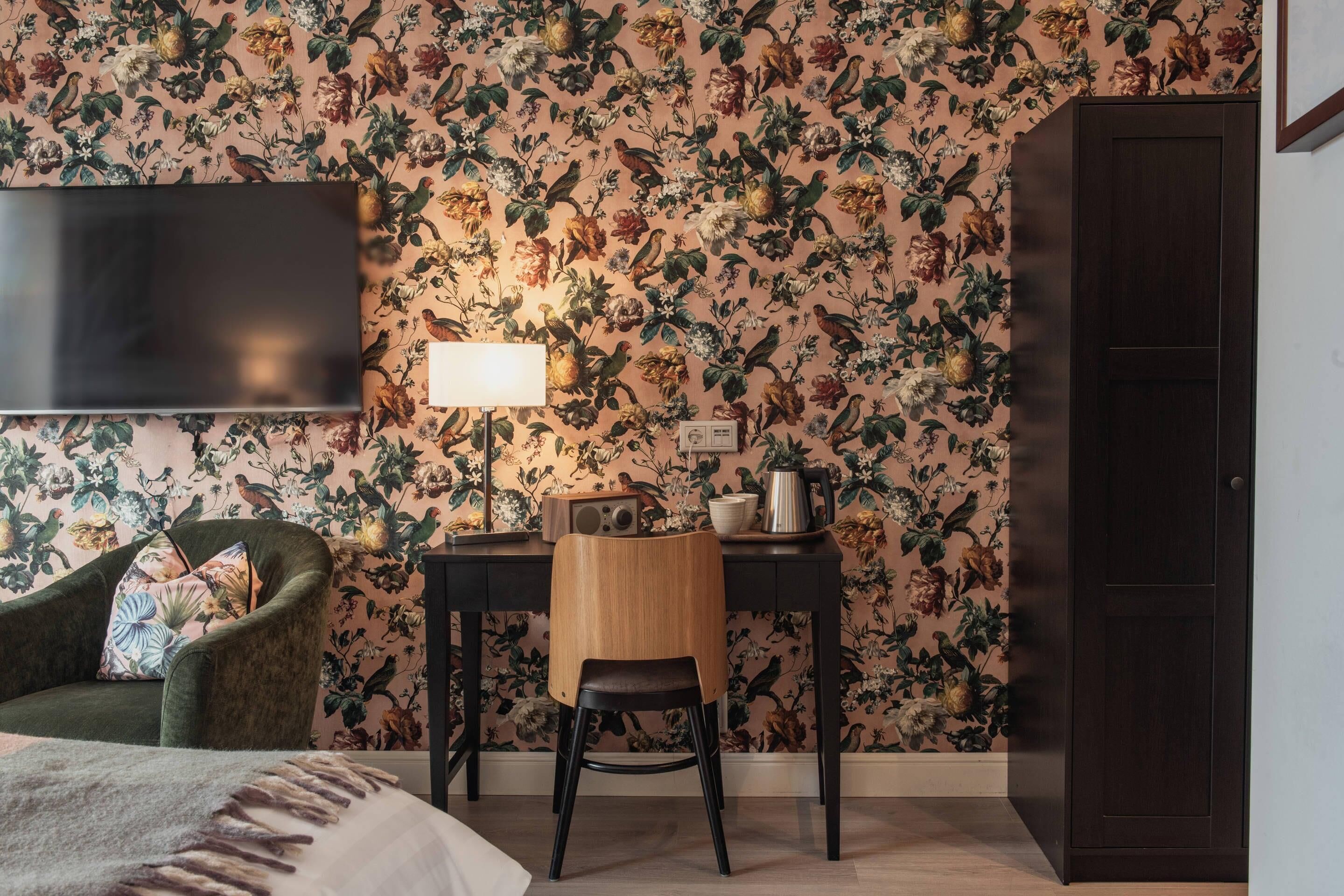 A room with a floral wallpaper, a desk with a lamp, a chair, a television, and a wardrobe.