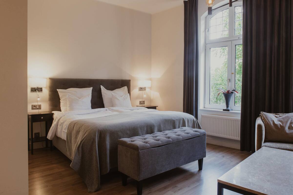 A bedroom with a bed, bedside tables, a bench, a window with curtains, and a view of greenery outside.