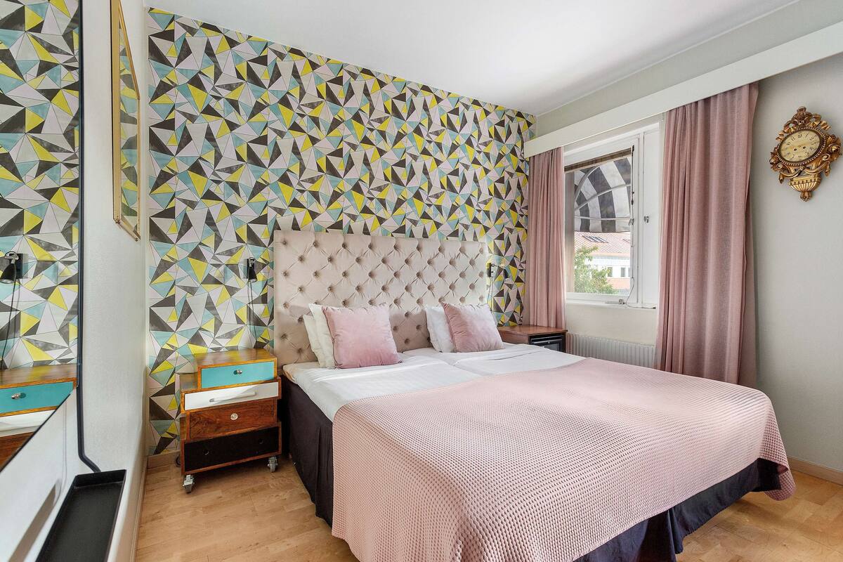 Une chambre avec un mur orné d’un motif géométrique, une tête de lit capitonnée, une couvre-lit rose et une fenêtre avec des rideaux.