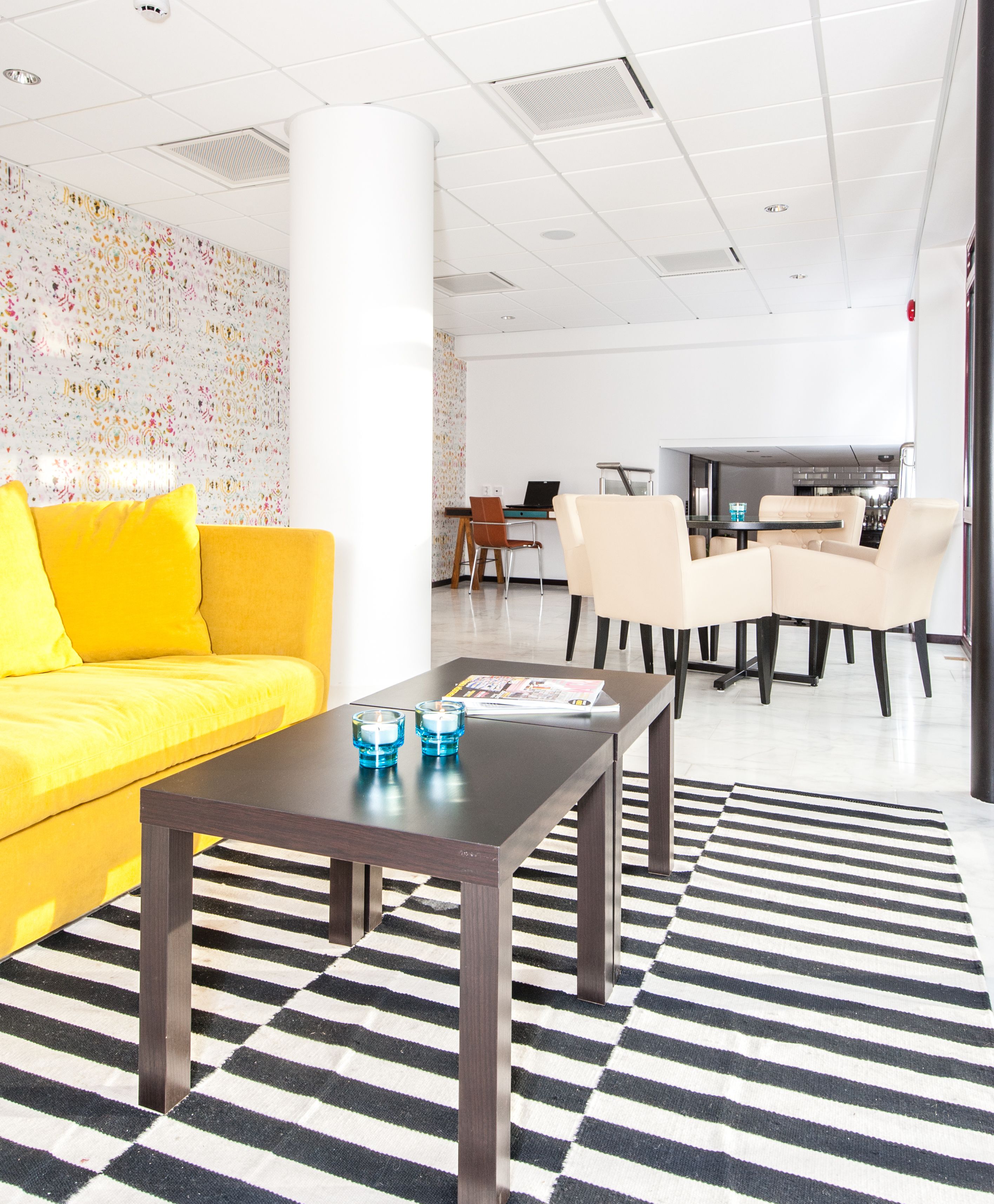 A modern lobby with a yellow sofa, a dark wooden coffee table, and a striped rug.