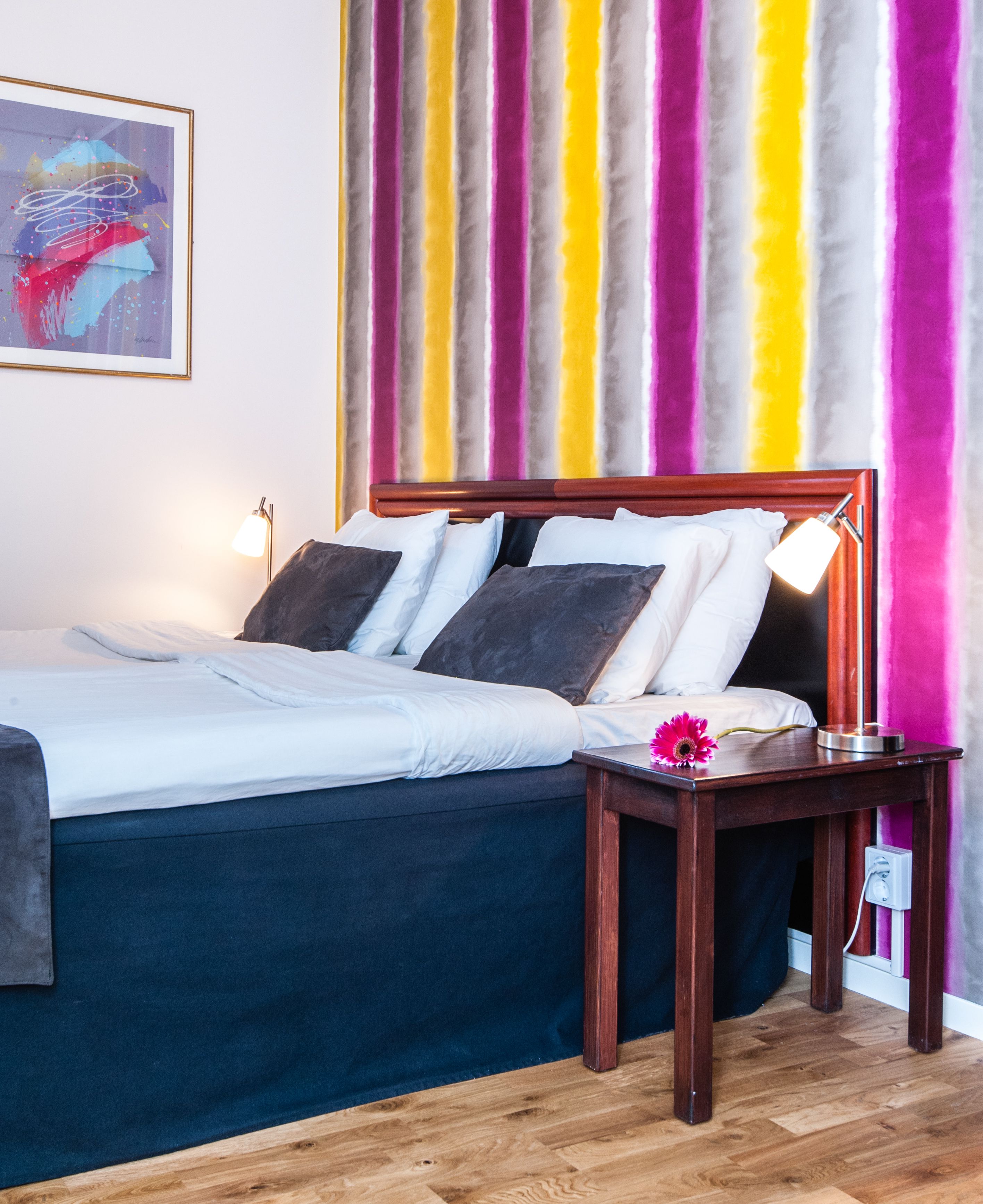 A neatly made bed with white and dark blue linens, a wooden nightstand with a lamp, and a vibrant striped curtain in the background.
