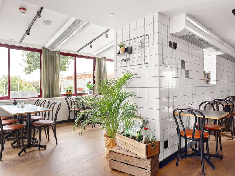 A restaurant with wooden tables and chairs, a large potted plant, and a tiled wall with a grid pattern.