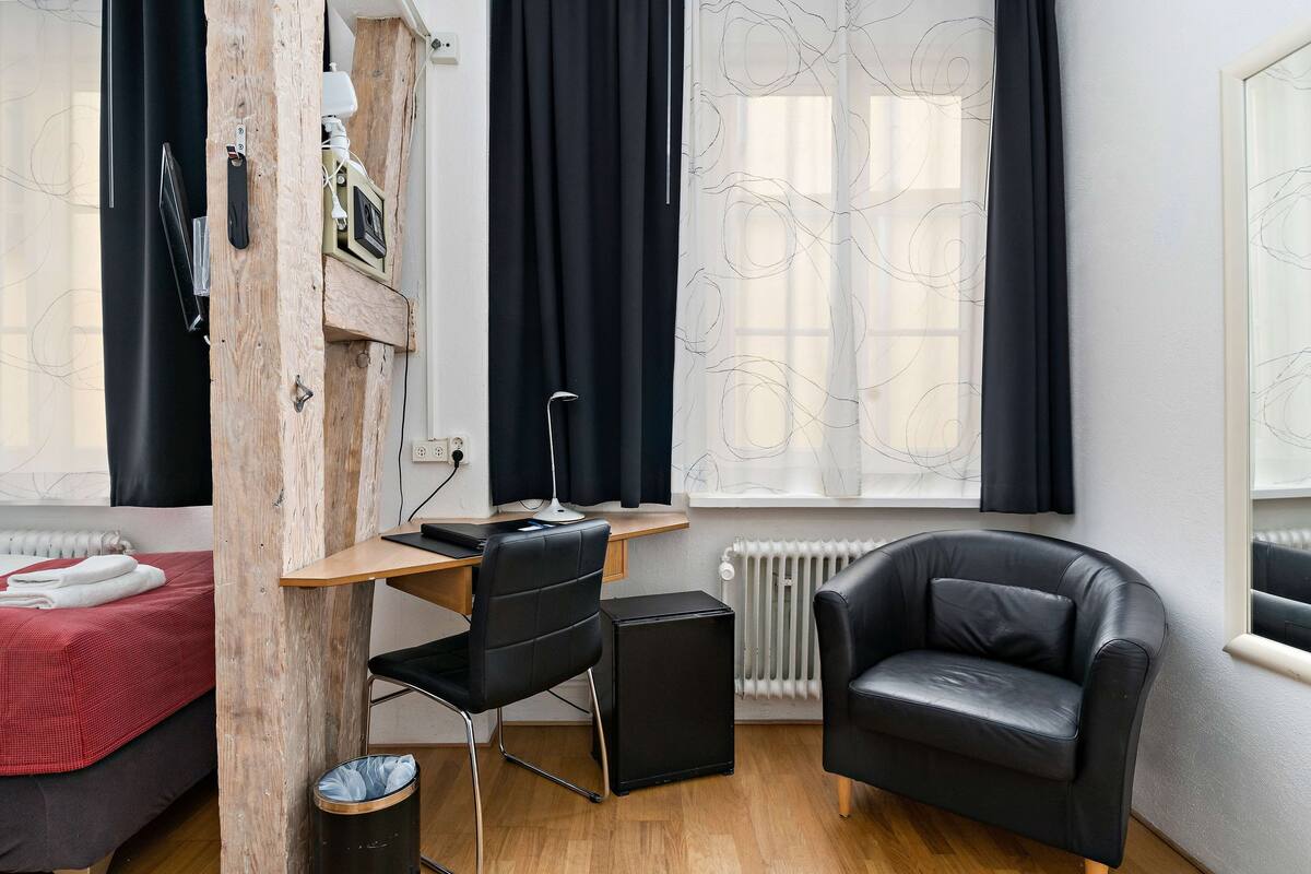 Quarto compacto com escrivaninha de madeira, cadeira preta, sofá de couro, radiador e janela com cortinas.