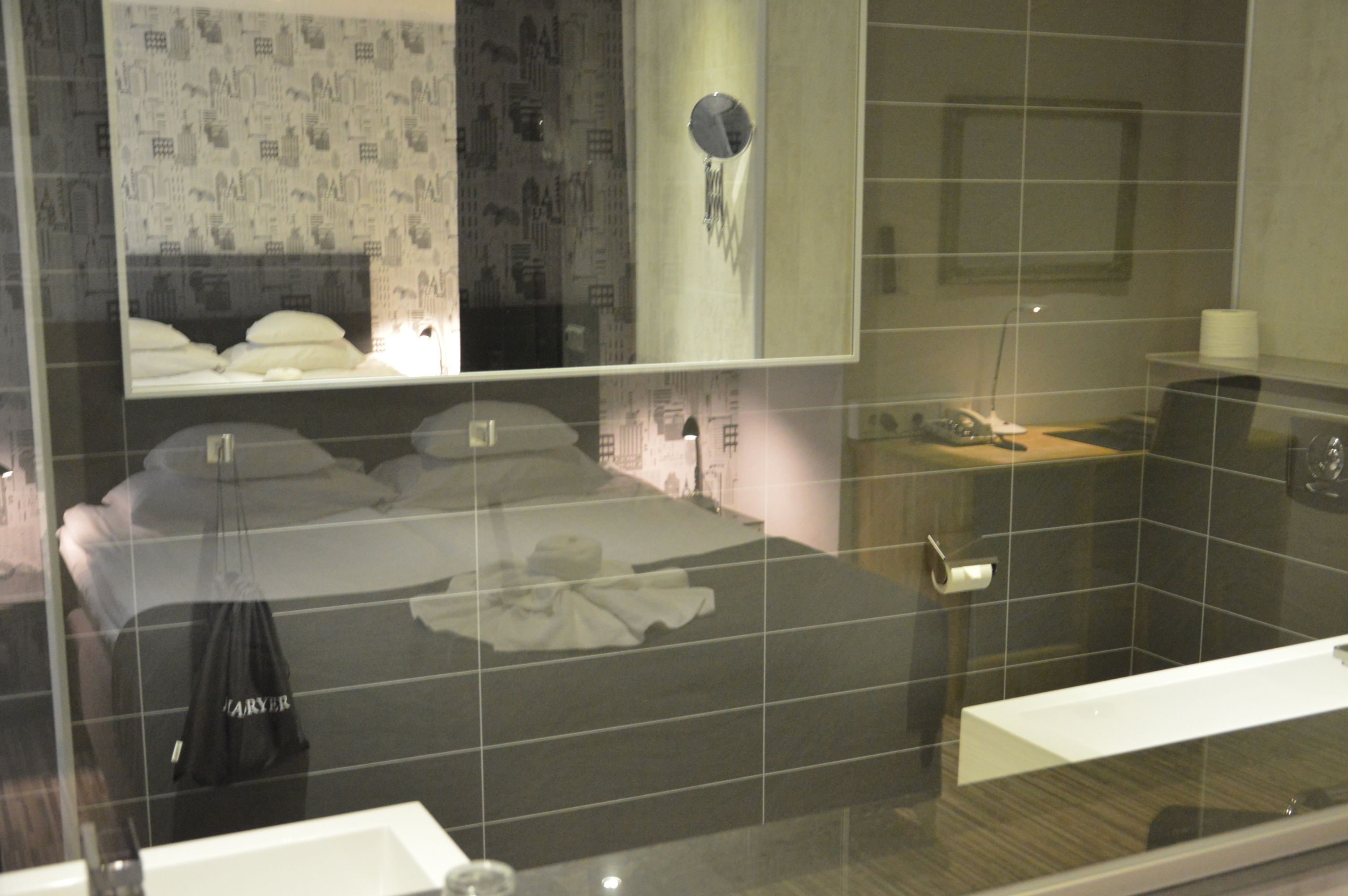 A hotel room with a large bed, a bedside table with a lamp, a mirror, and a bathroom with a sink and toilet.