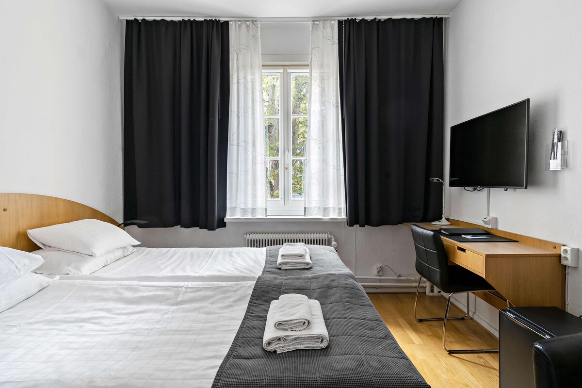 A hotel room with two beds, a desk, a chair, a TV, and a window with curtains.