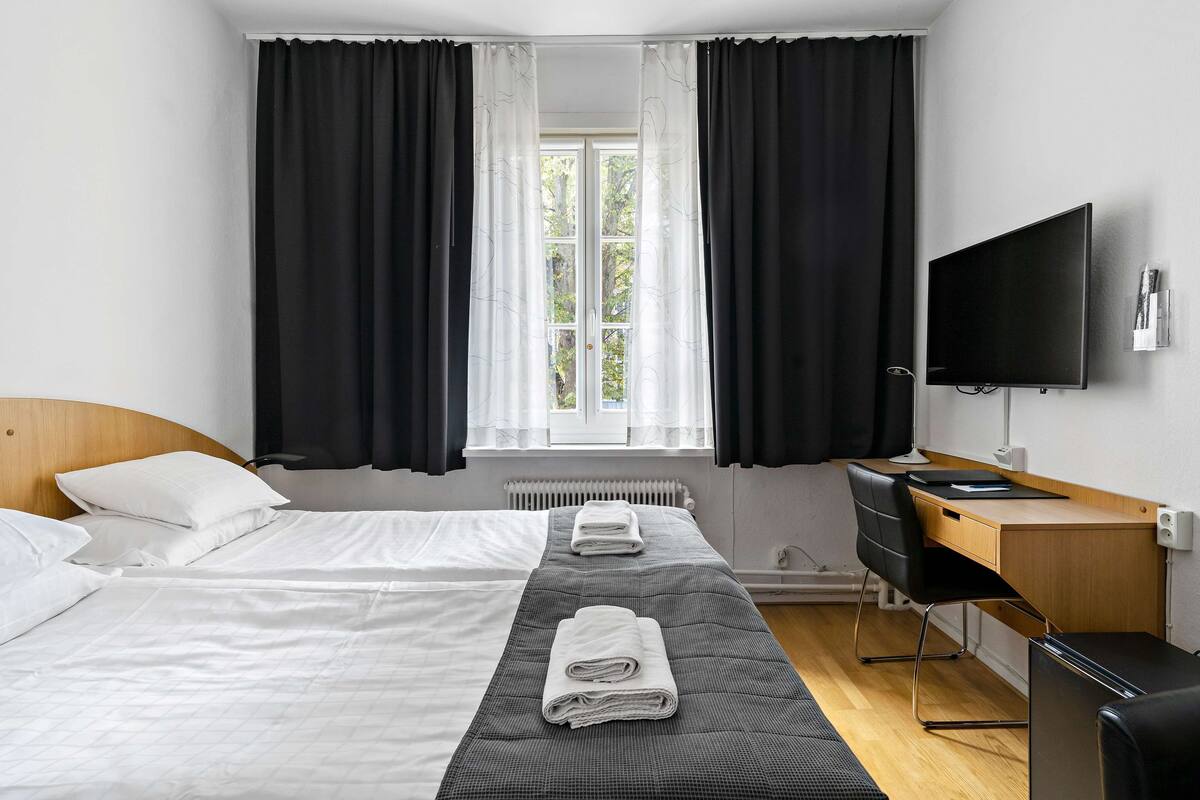 Quarto de hotel com duas camas, uma escrivaninha, uma cadeira, uma televisão e uma janela com cortinas.