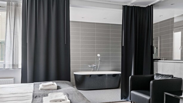 Habitación de hotel moderna con una cama grande, un sillón de cuero negro, una bañera y un ventanal con cortinas.
