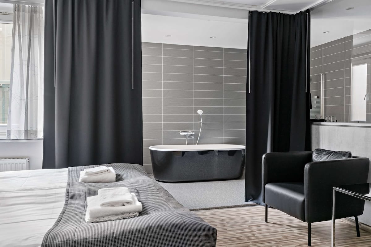 Quarto de hotel moderno com uma cama grande, uma poltrona de couro preto, banheira e uma janela ampla com cortinas.