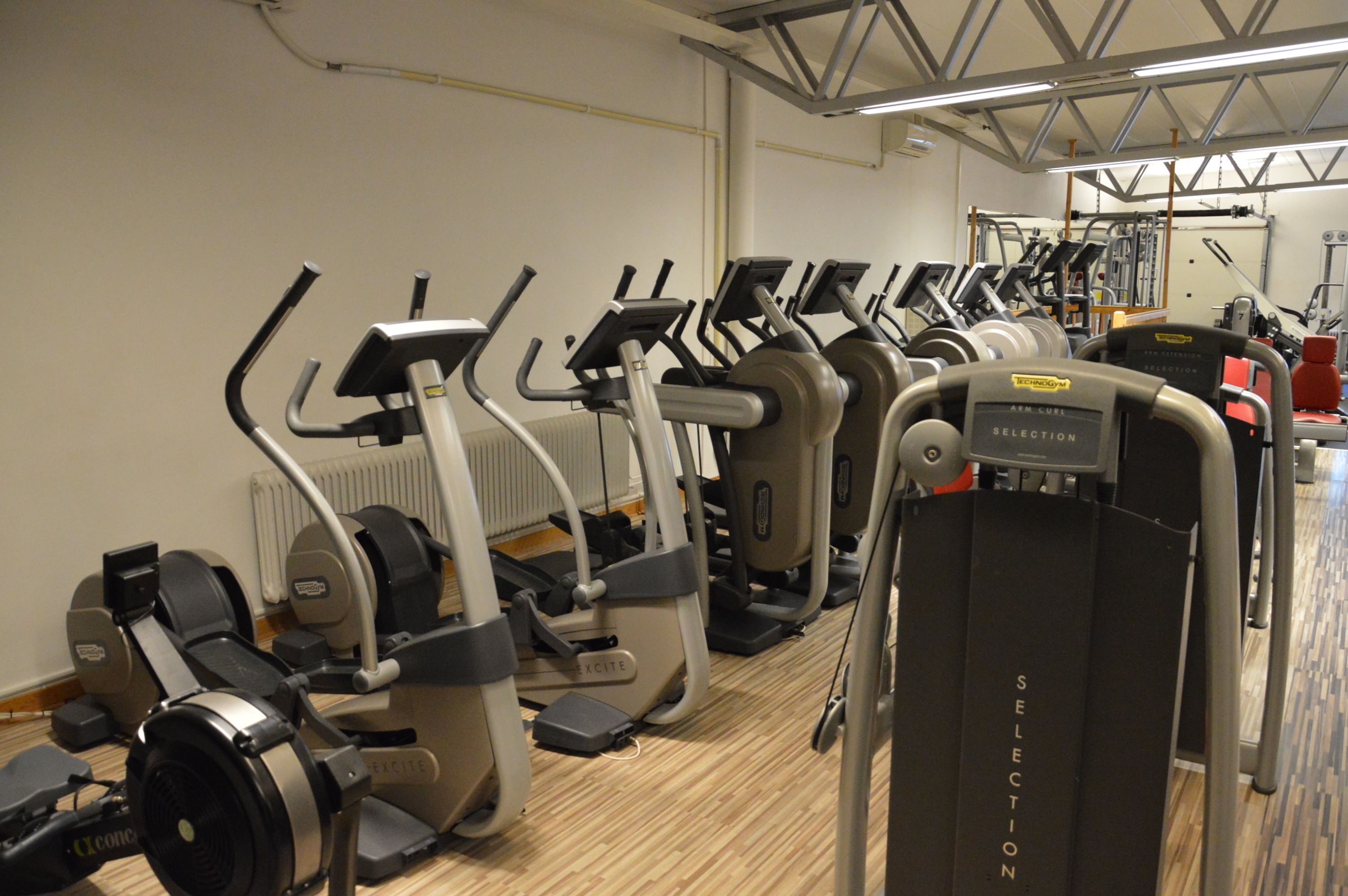 En rad stillastående träningsmaskiner i ett gym med trägolv och vita väggar.