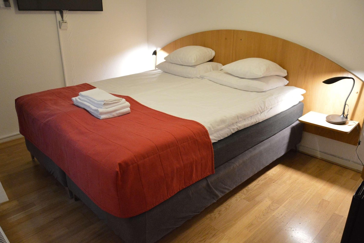 A neatly made bed with a red blanket, white pillows, and a grey mattress. There is a bedside table with a lamp and a power outlet.