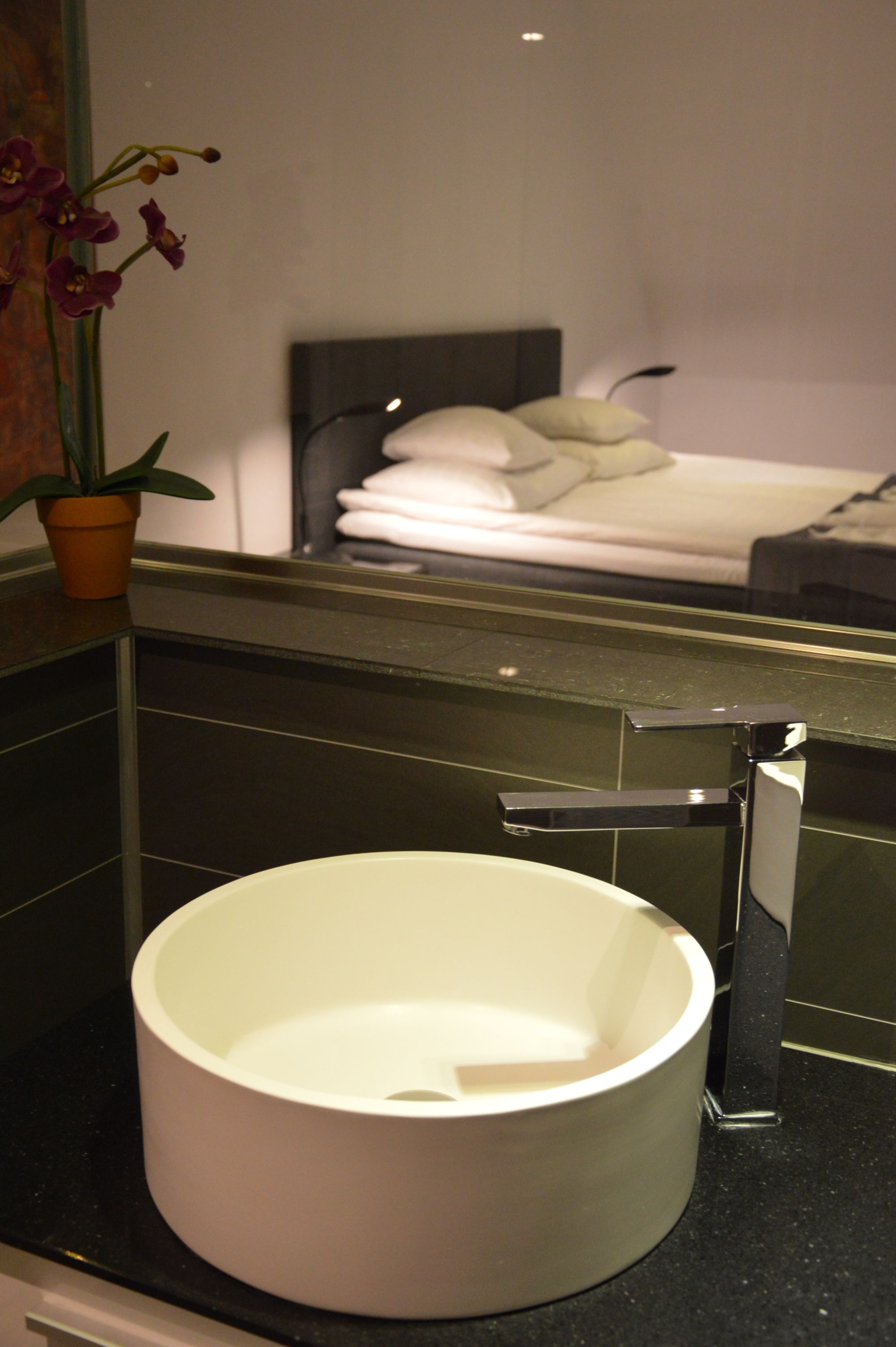 Une salle de bain avec un lavabo blanc, un miroir et un lit en arrière-plan.