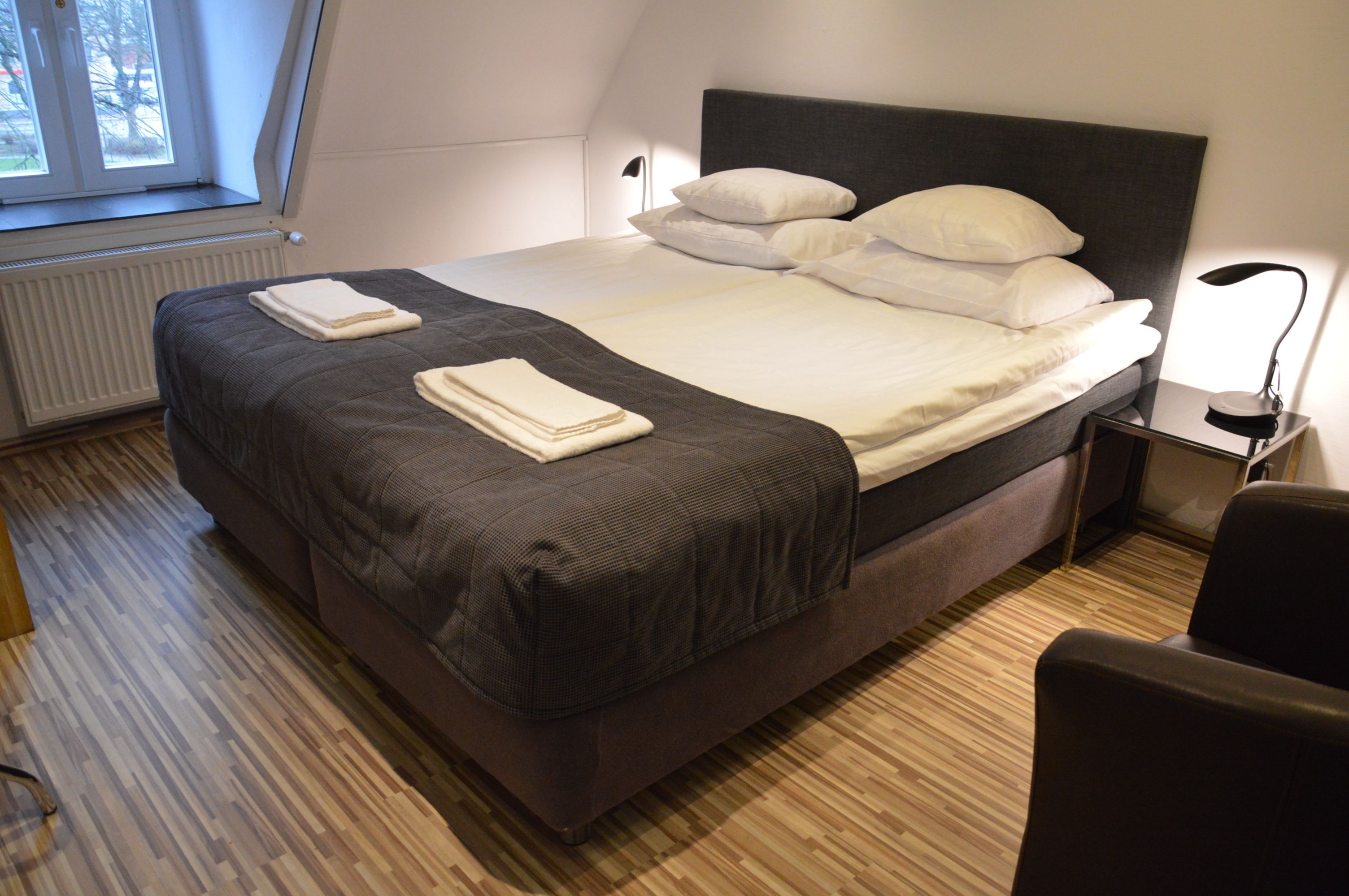 A neatly made bed with a dark headboard and white linens, a bedside lamp, and a view of the outdoors through a window.