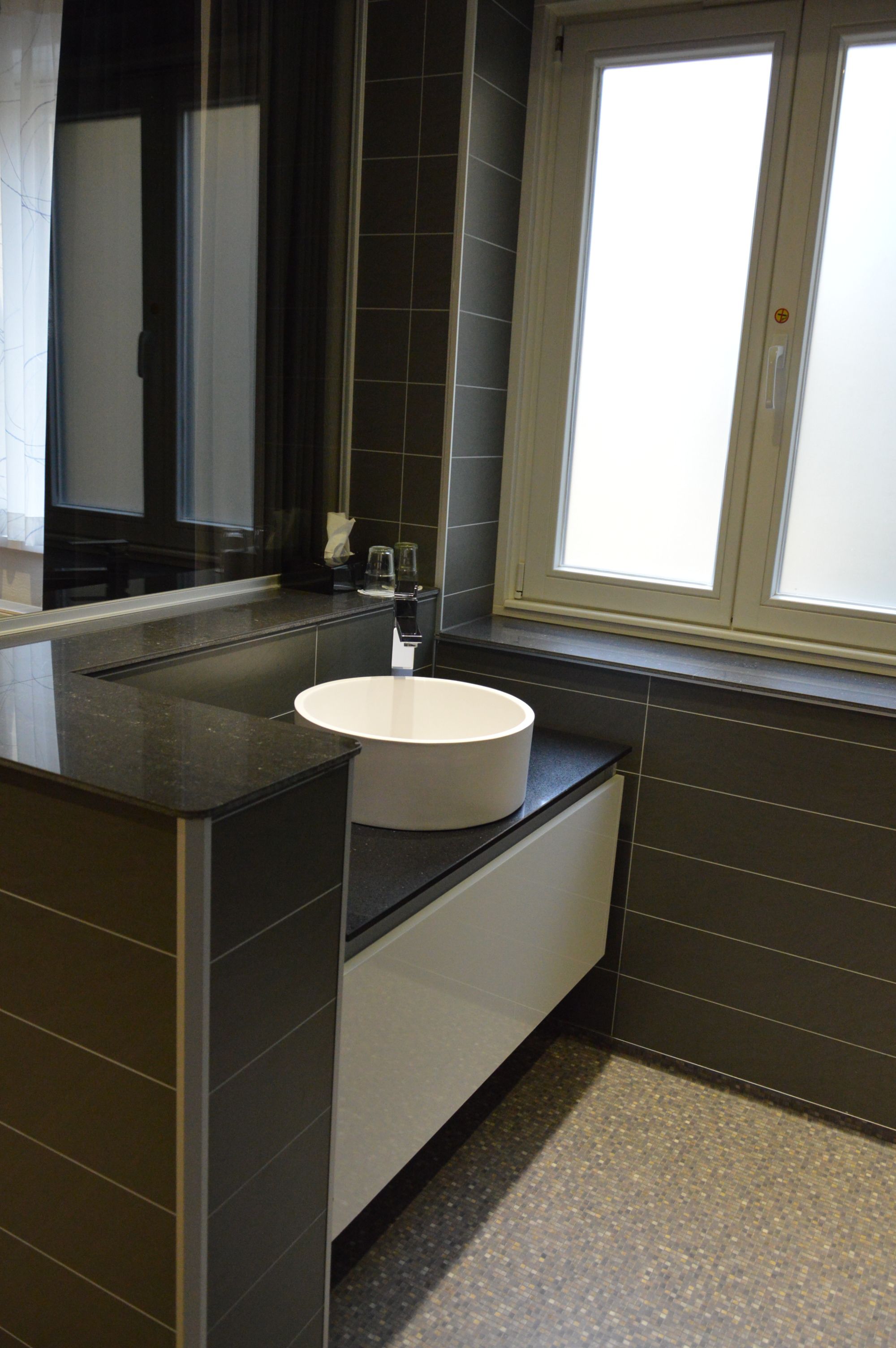 A modern bathroom with a white sink, dark tiles, and a window providing natural light.