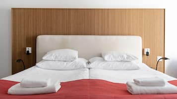 Una cama bien hecha con sábanas blancas y una manta roja, un cabecero de madera y dos mesitas de noche fijadas a la pared.