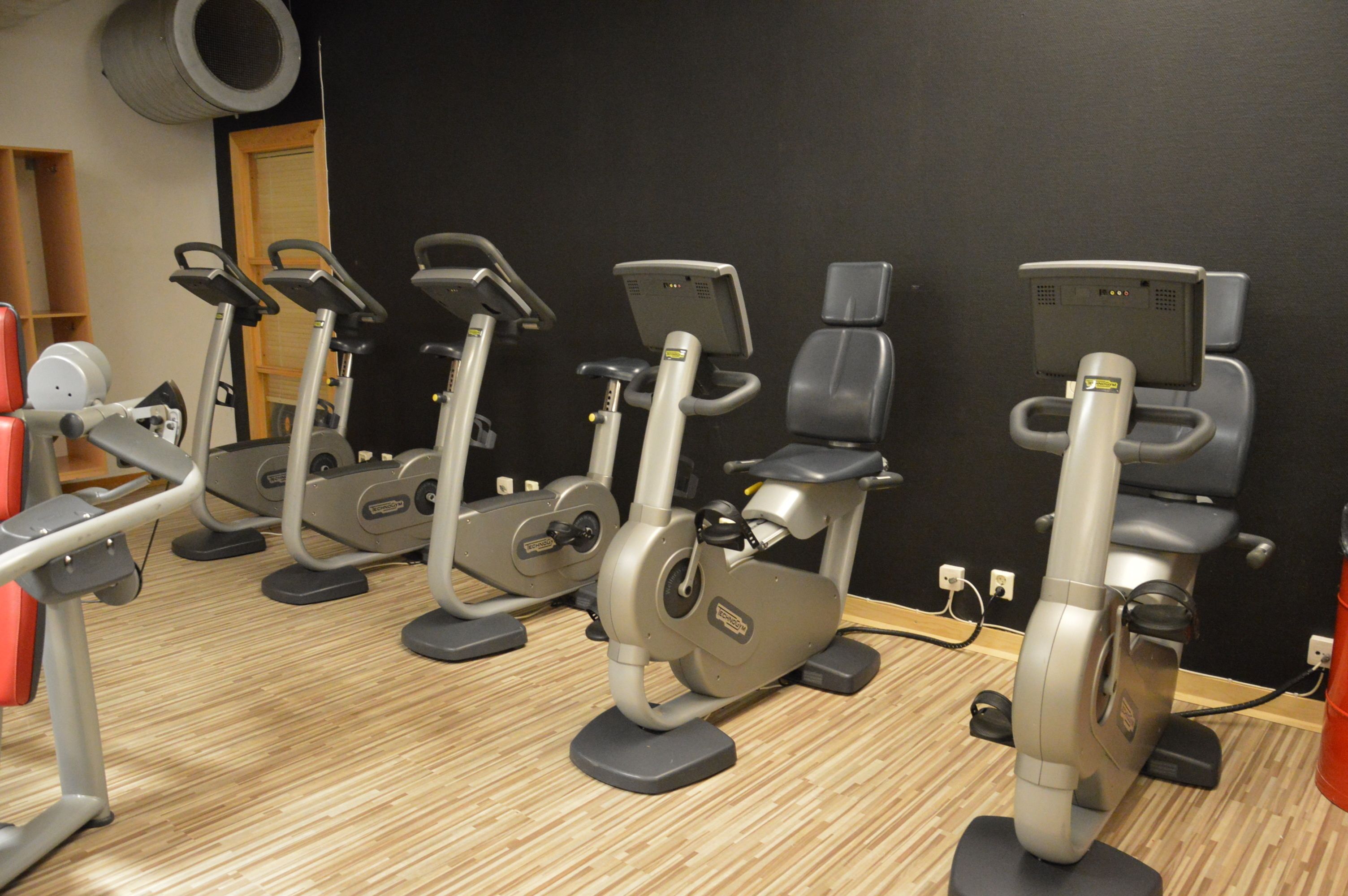 Række stillestående træningscykler i et fitnesscenter med trægulv og sort væg.