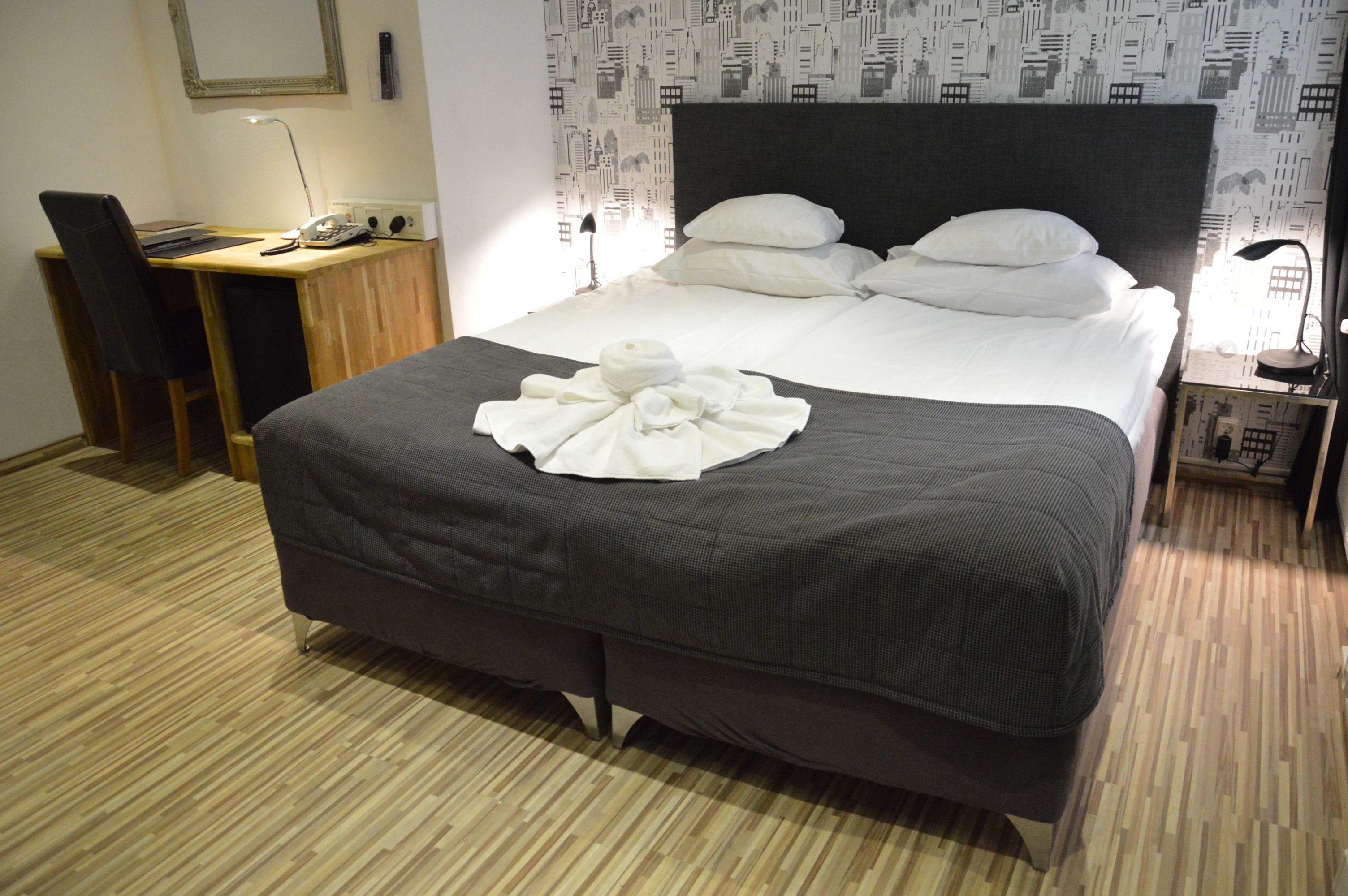 A neatly made bed with a dark headboard, a wooden desk with a chair, and a lamp on a nightstand.