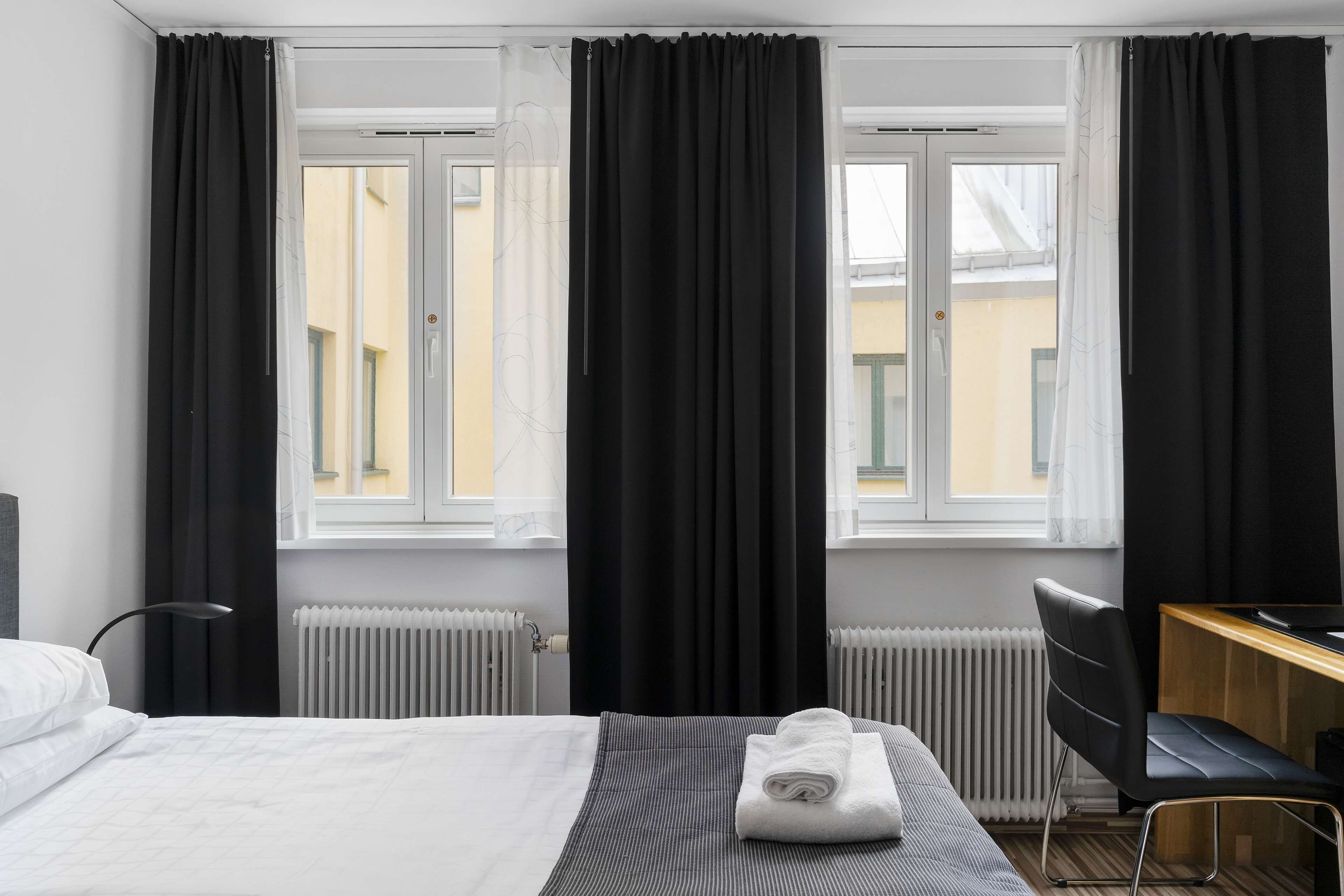 Ett hotellrum med en säng, ett skrivbord och en stol. Det finns två fönster med mörka gardiner, ett element och en lampa.