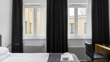 Ett hotellrum med en säng, ett skrivbord och en stol. Det finns två fönster med mörka gardiner, ett element och en lampa.