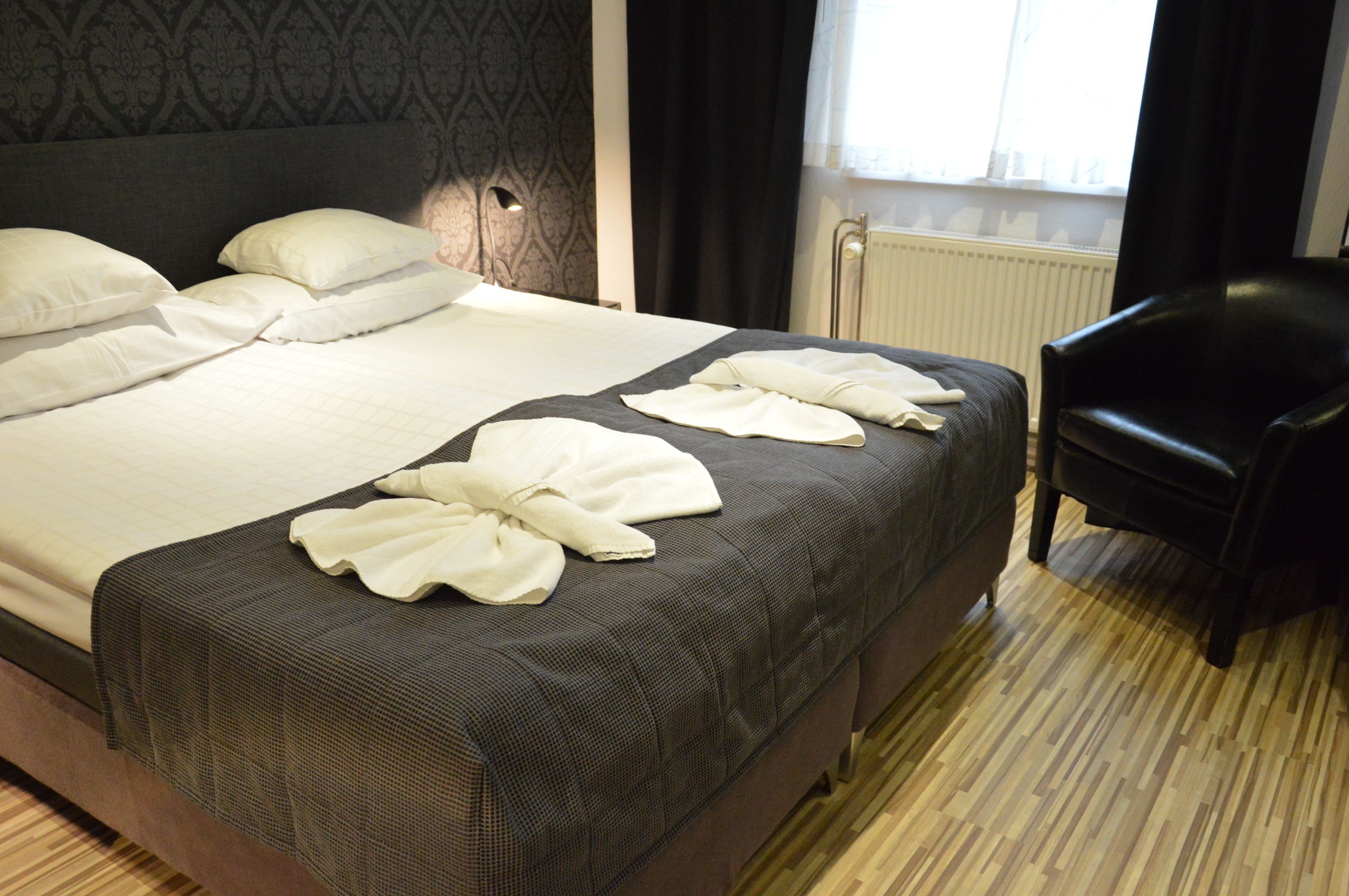 A neatly made bed with white linens and towels, a dark patterned headboard, and a wooden floor.