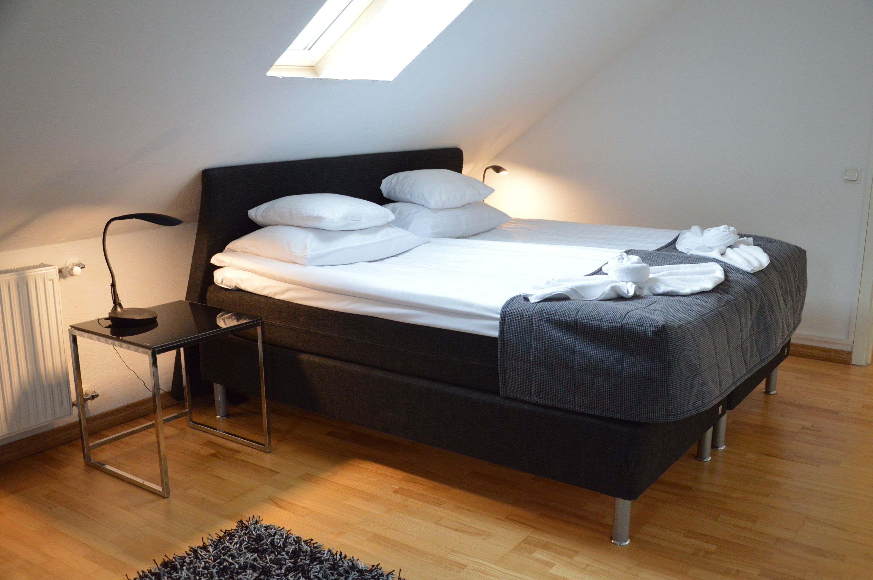 Un dormitorio moderno con tragaluz, una cama con ropa de cama blanca, una mesita de noche con lámpara y una pequeña alfombra.