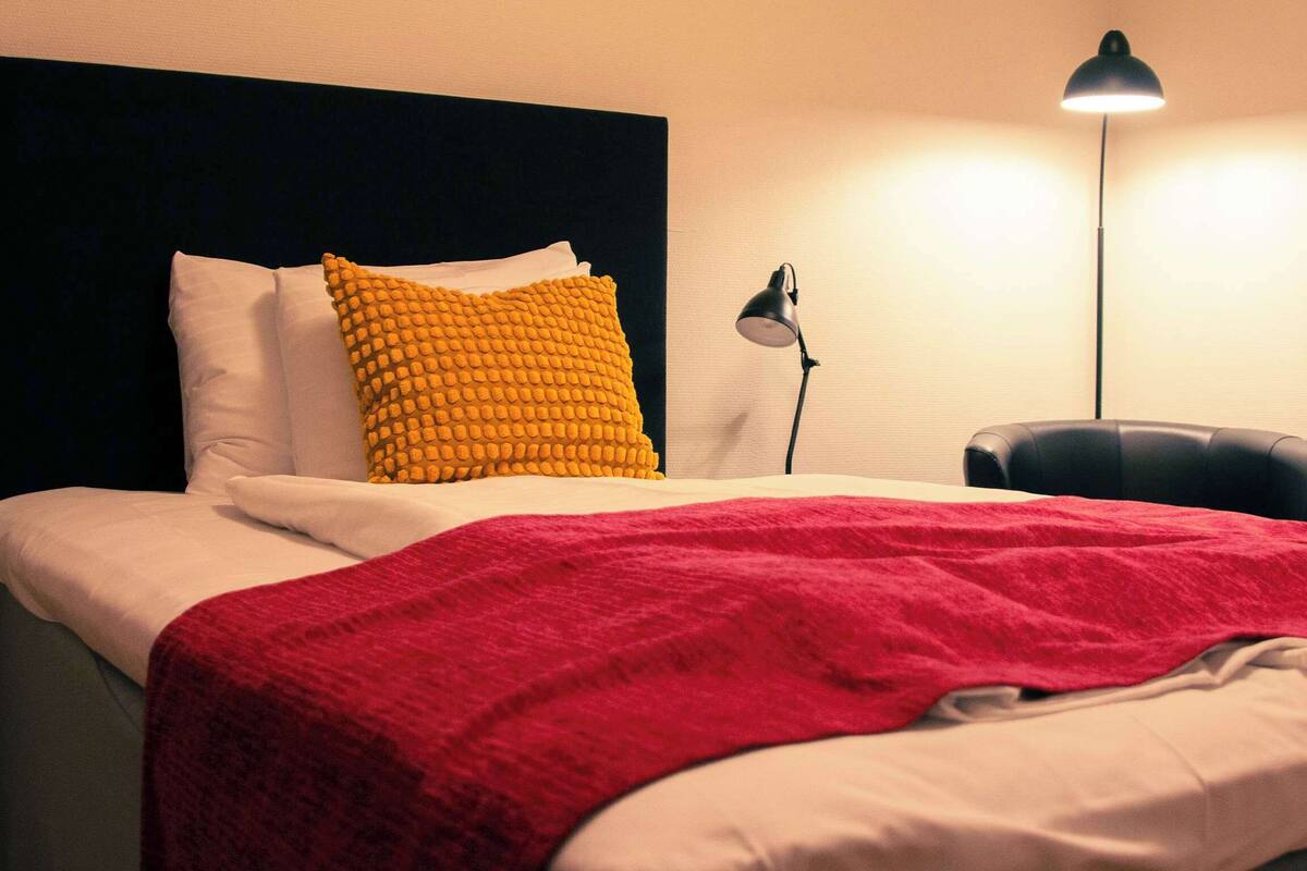 A neatly made bed with white and red bedding, a black headboard, and a yellow textured pillow.