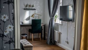 A room with a wooden floor, a desk with a lamp, a chair, a television mounted on the wall, and a window with curtains.