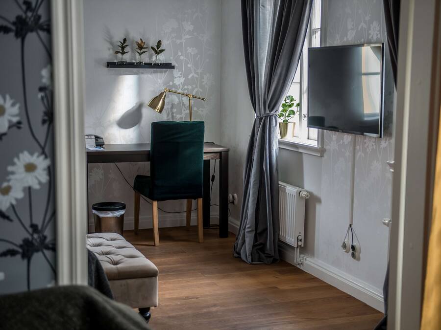 A room with a wooden floor, a desk with a lamp, a chair, a television mounted on the wall, and a window with curtains.