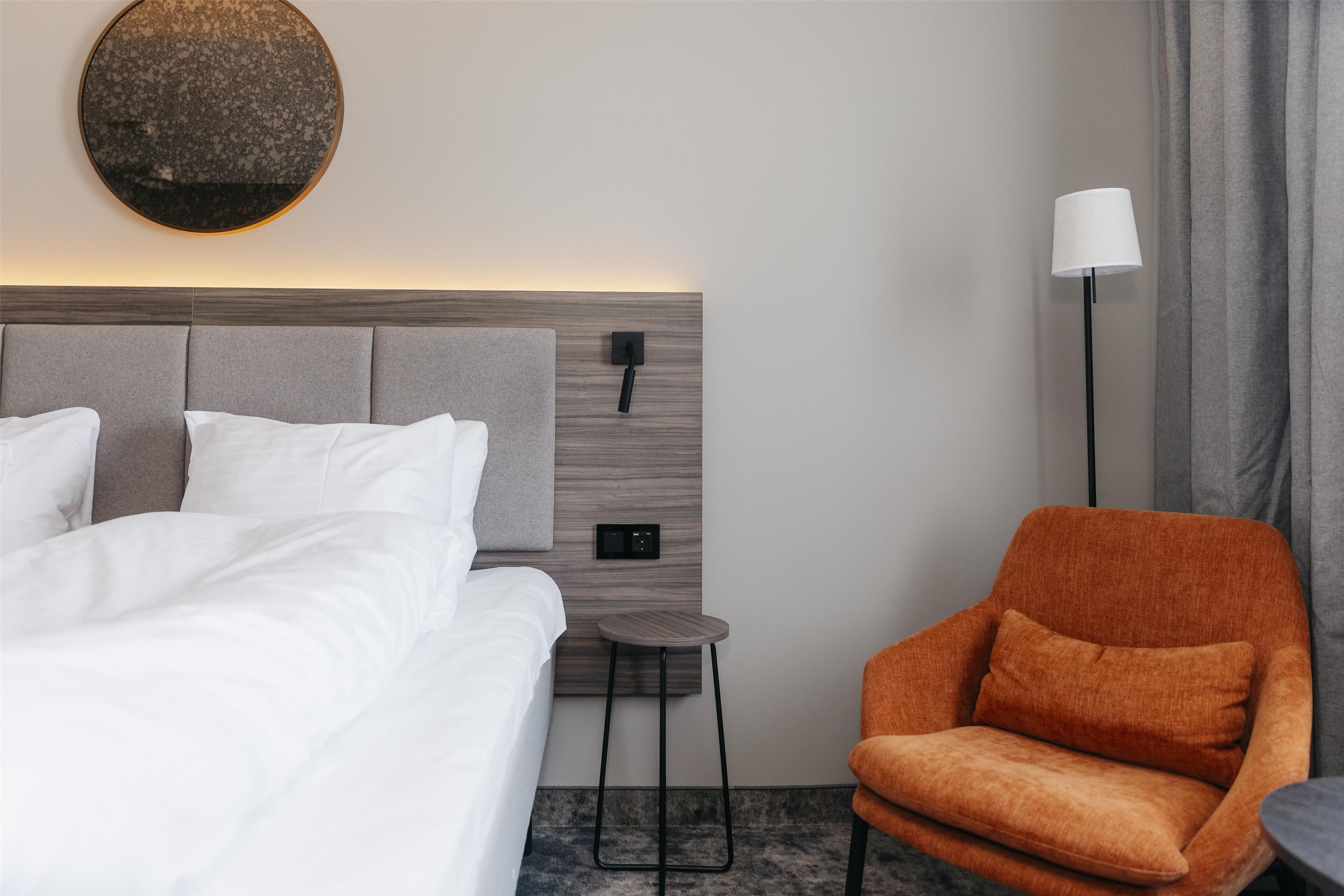 Habitación de hotel con una cama, un espejo redondo, una mesita, una lámpara y una silla naranja.