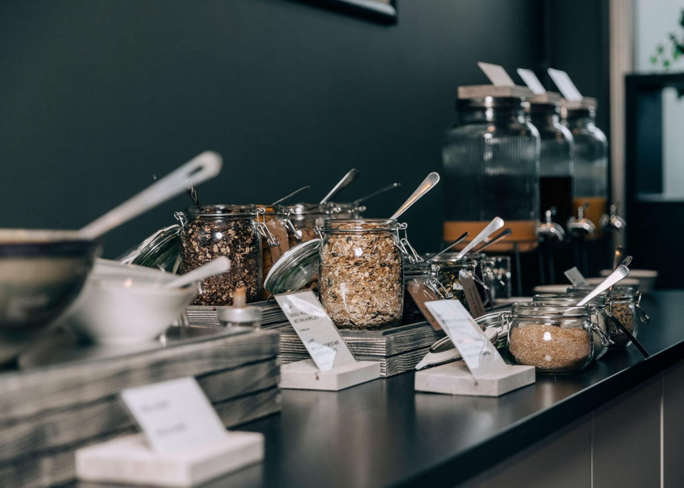 A self-serve station with various food items in jars, each with a spoon and price tag.