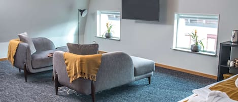 A modern hotel room with a large bed, two armchairs, a floor lamp, a TV, and two windows with potted plants.