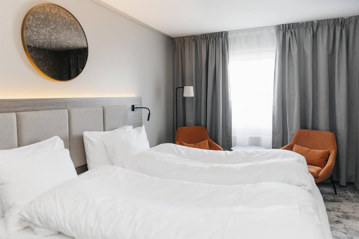 A hotel room with a large bed, two chairs, a round mirror, a lamp, and a window with curtains.