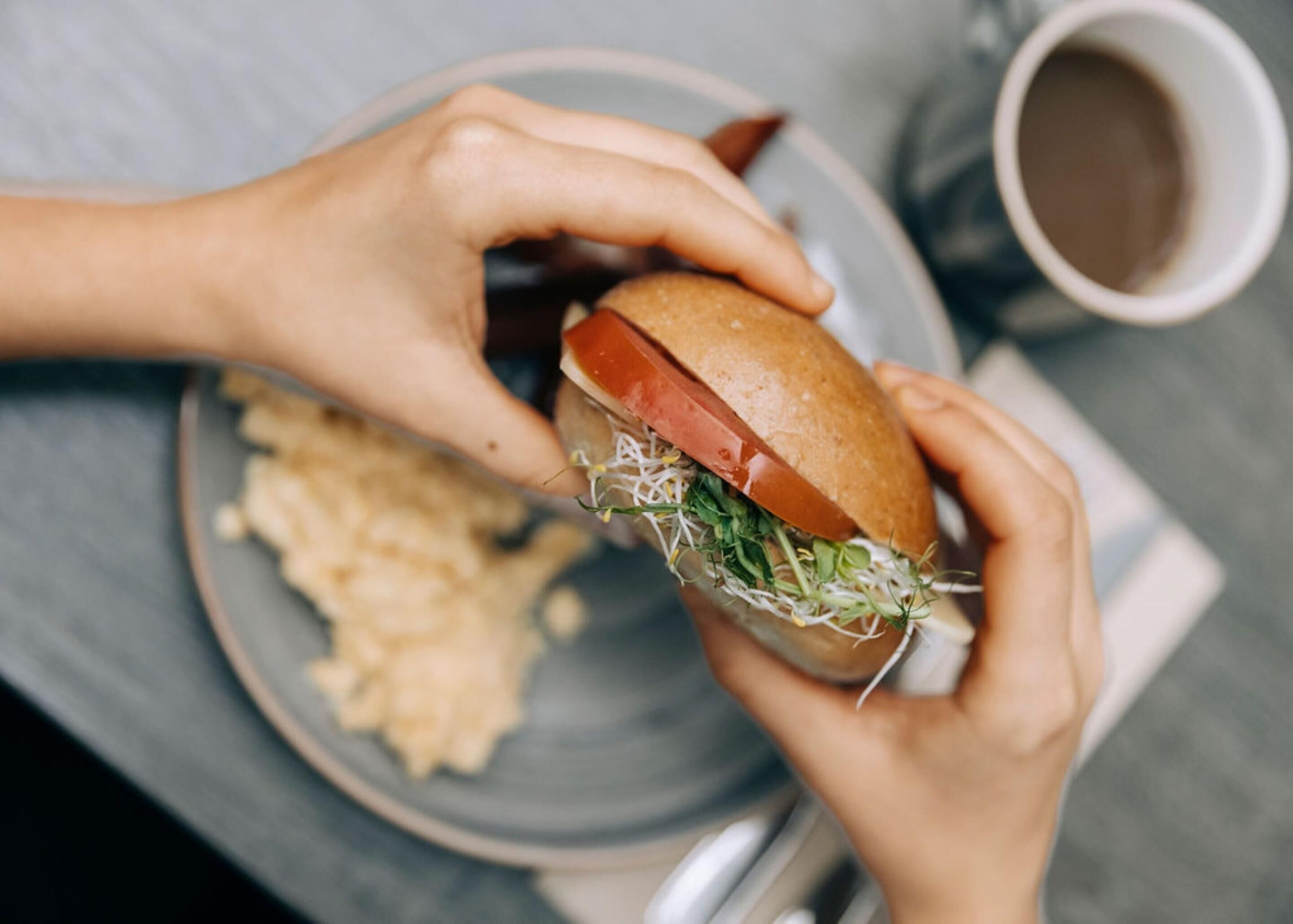 A person holding a sandwich with a bun, tomato, and greens, and a plate of chips.