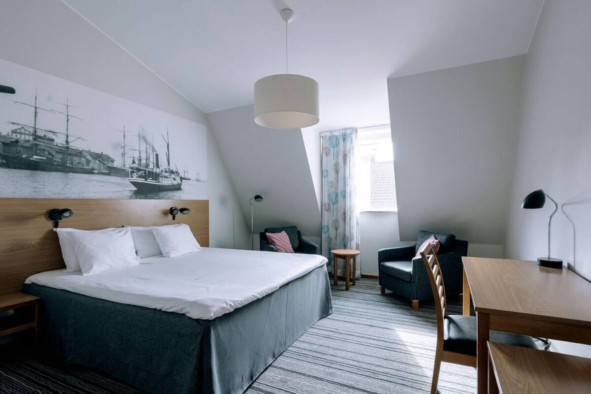 A modern hotel room with a large bed, a desk, a chair, a small table, and a ceiling light.