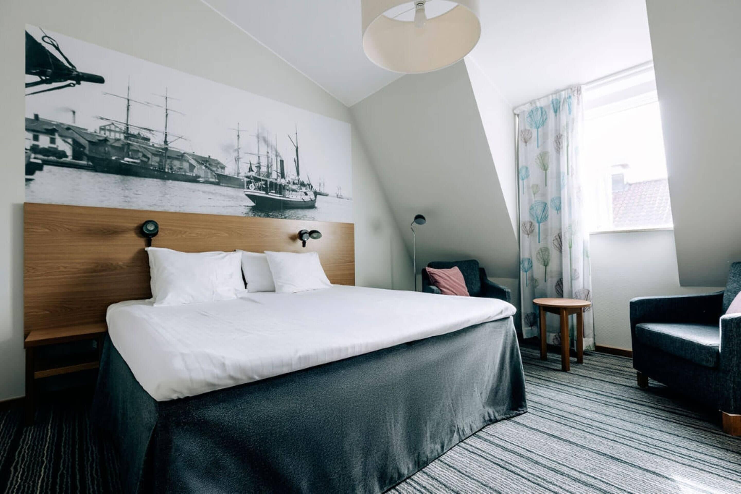 A hotel room with a large bed, a headboard with a wooden panel, a wall-mounted lamp, a small round table, a chair, and a window with curtains.