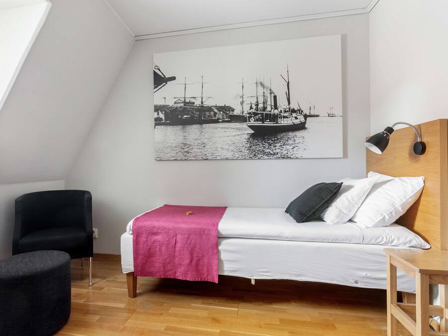 A bedroom with a bed, a chair, a small table, a wall-mounted lamp, and a large black and white maritime painting.