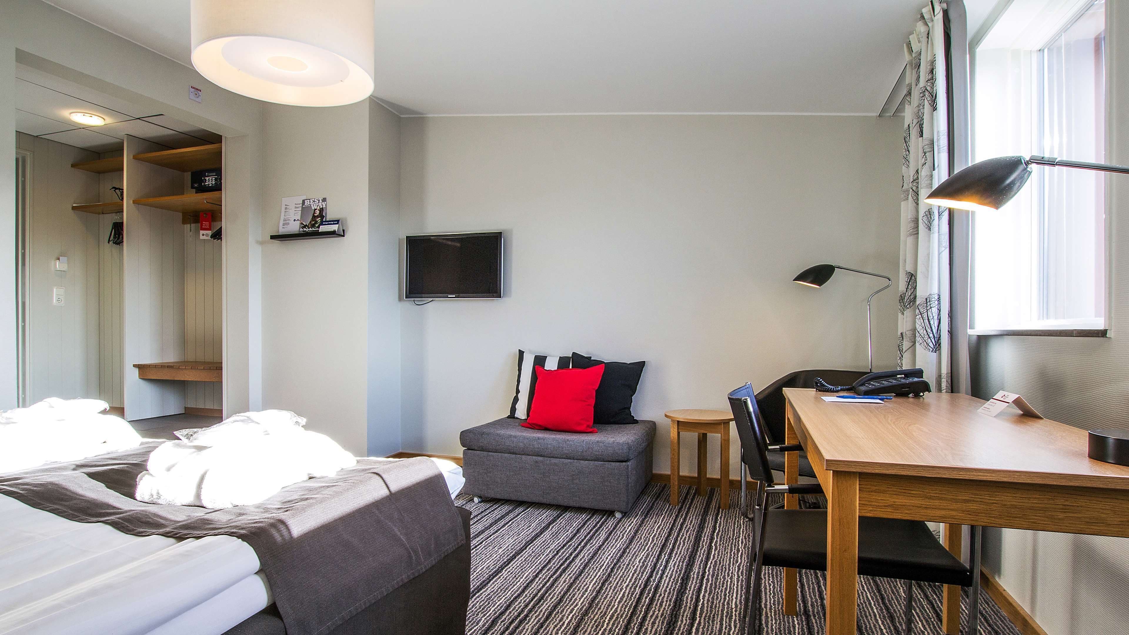 Habitación de hotel compacta con cama, escritorio, silla, un pequeño sofá, televisor y ventana con cortinas.