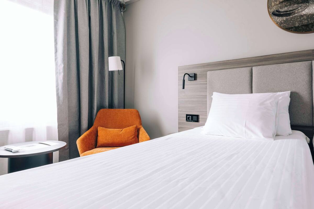 A hotel room with a large bed, a small round table, an orange armchair, and a wall-mounted lamp.