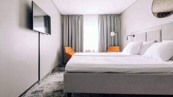 A hotel room with a bed, two chairs, a TV, a lamp, and a window with curtains.