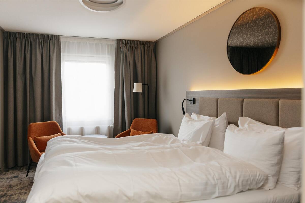 A hotel room with a large bed, two chairs, a round mirror, and a window with curtains.