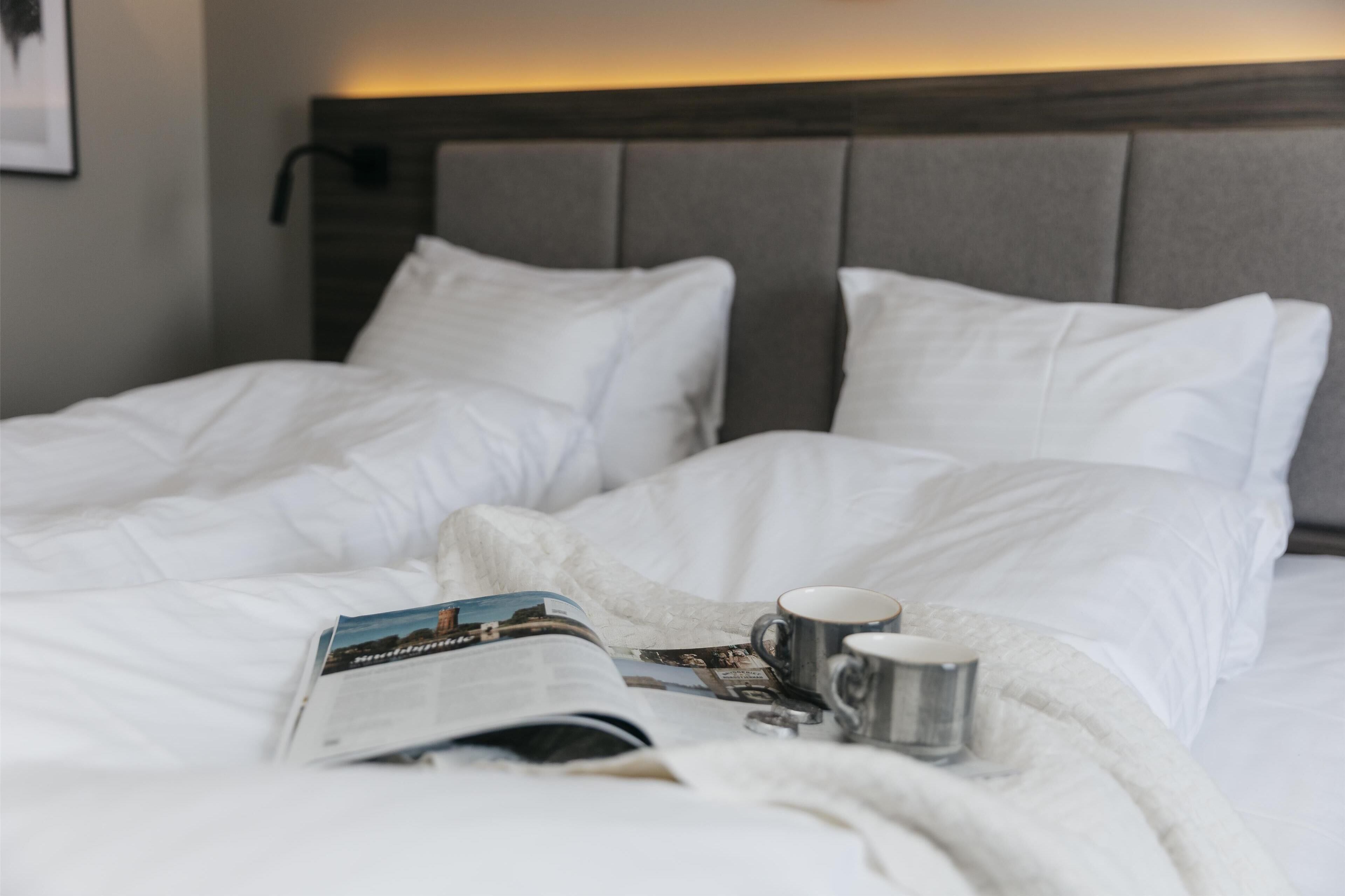 A neatly made bed with white linens, a magazine, and a cup on the bed.