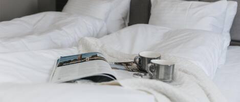 Una cama bien tendida con sábanas blancas, una revista y una taza sobre la cama.